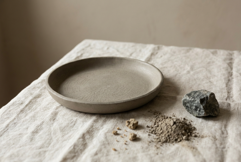 Shallow ceramic dish of smooth grey-beige mineral healing clay with a finely cracked surface on warm cream linen in soft window light, a rough mineral stone beside it.